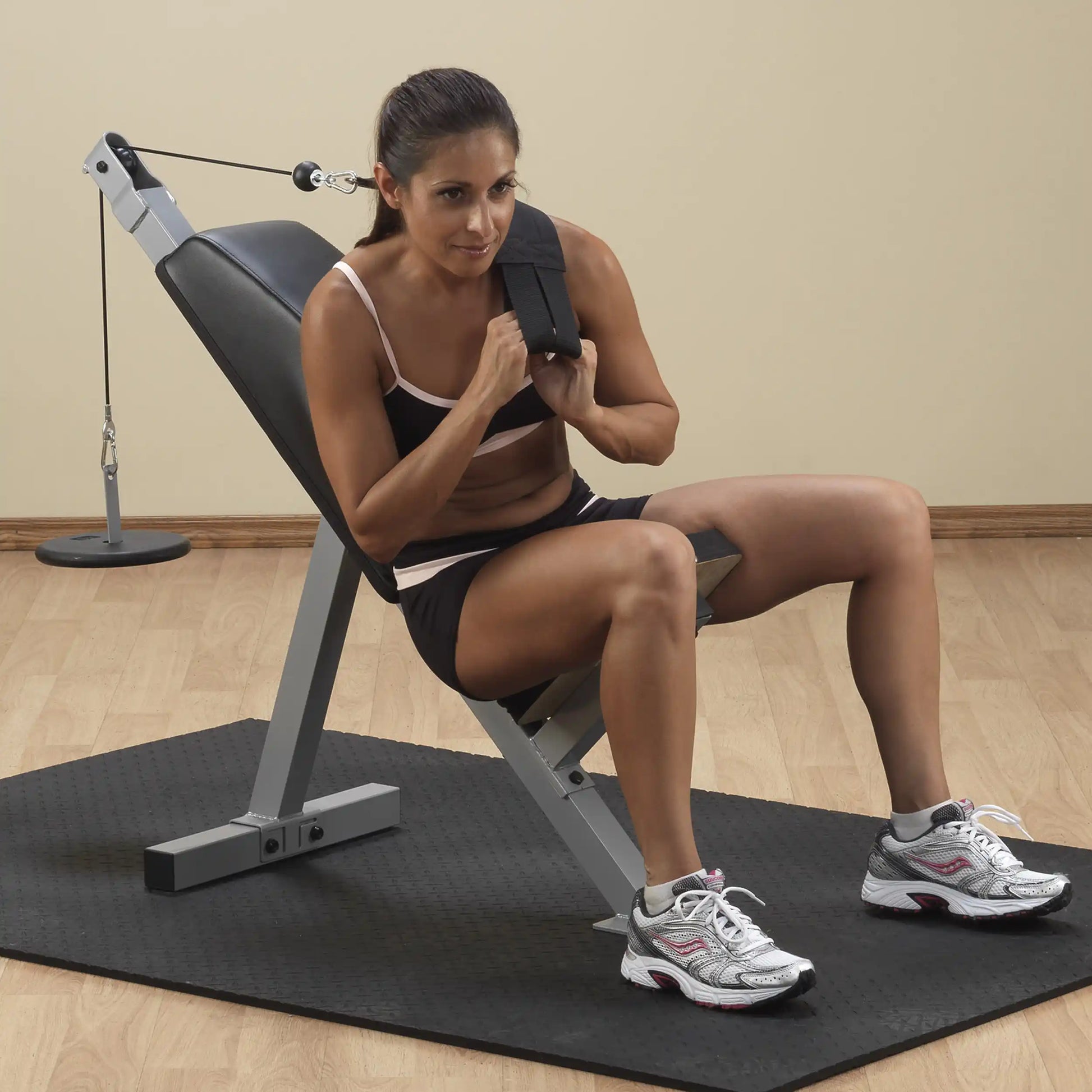 A woman in athletic clothing sitting on an adjustable workout bench, using a resistance cable for strength training. She is focused and positioned for exercise, with a wooden floor and a gym setup in the background.