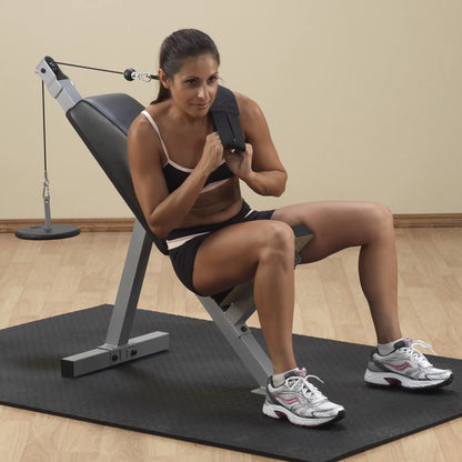 A woman in athletic clothing sitting on an adjustable workout bench, using a resistance cable for strength training. She is focused and positioned for exercise, with a wooden floor and a gym setup in the background.