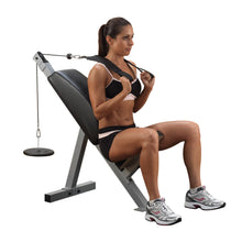 A woman sitting on a weight bench, performing a seated cable row exercise. She is wearing a black sports bra and shorts, with her hair tied back. The exercise equipment includes a cable attached to a weight plate.