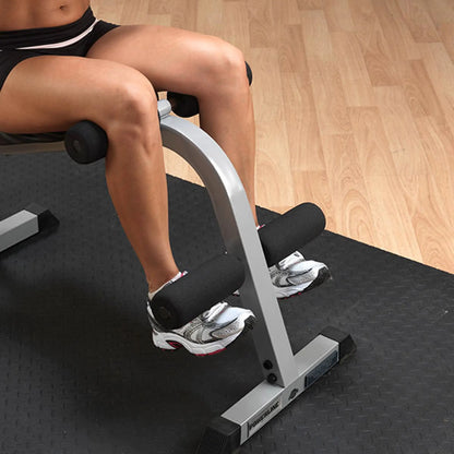 Close-up of a person's legs positioned on a workout machine, demonstrating a leg exercise, with focus on the padded supports and athletic shoes on a textured gym floor.