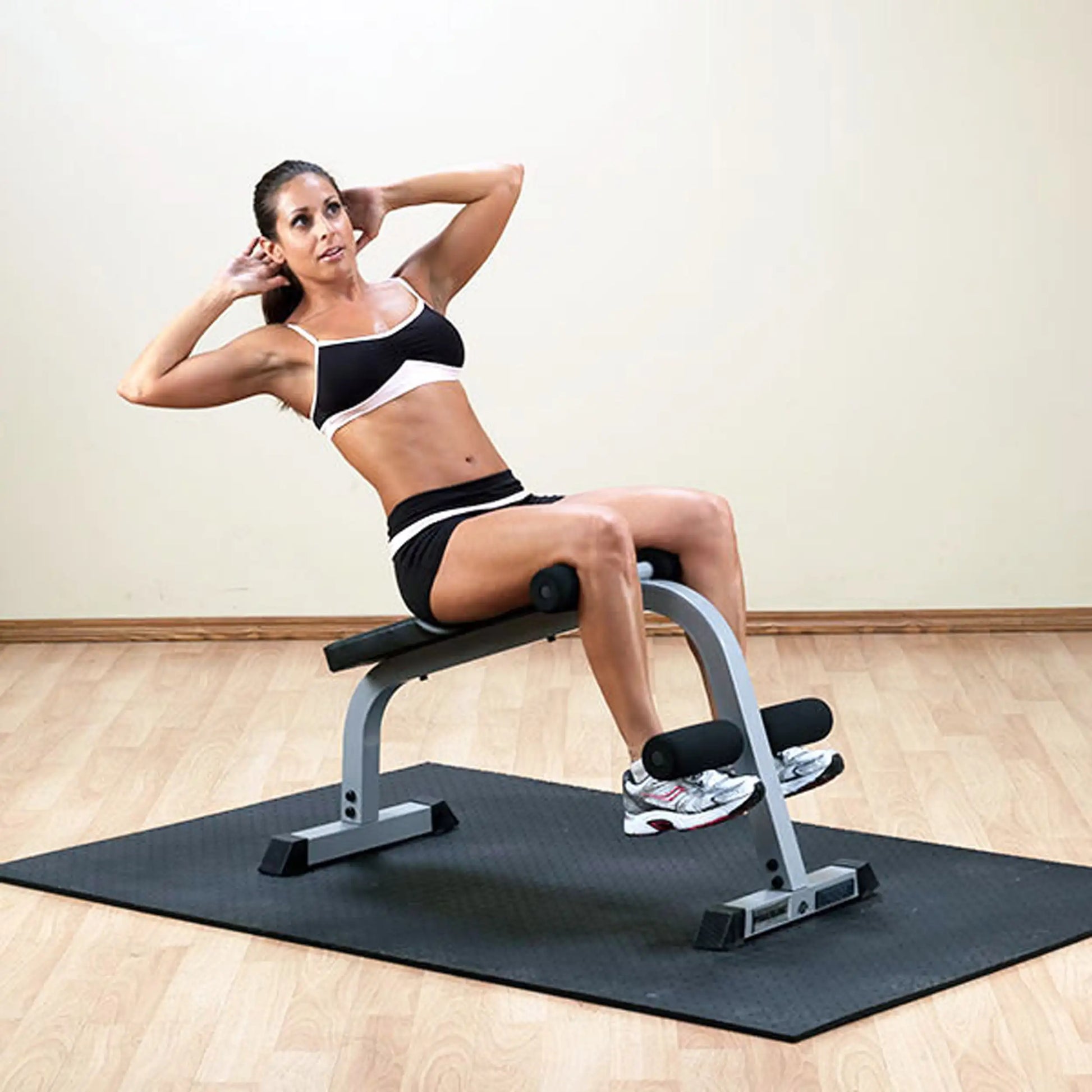 A woman performing abdominal exercises on a workout bench, wearing a black and white sports outfit, with her hands behind her head and legs bent, on a rubber mat in a fitness studio.