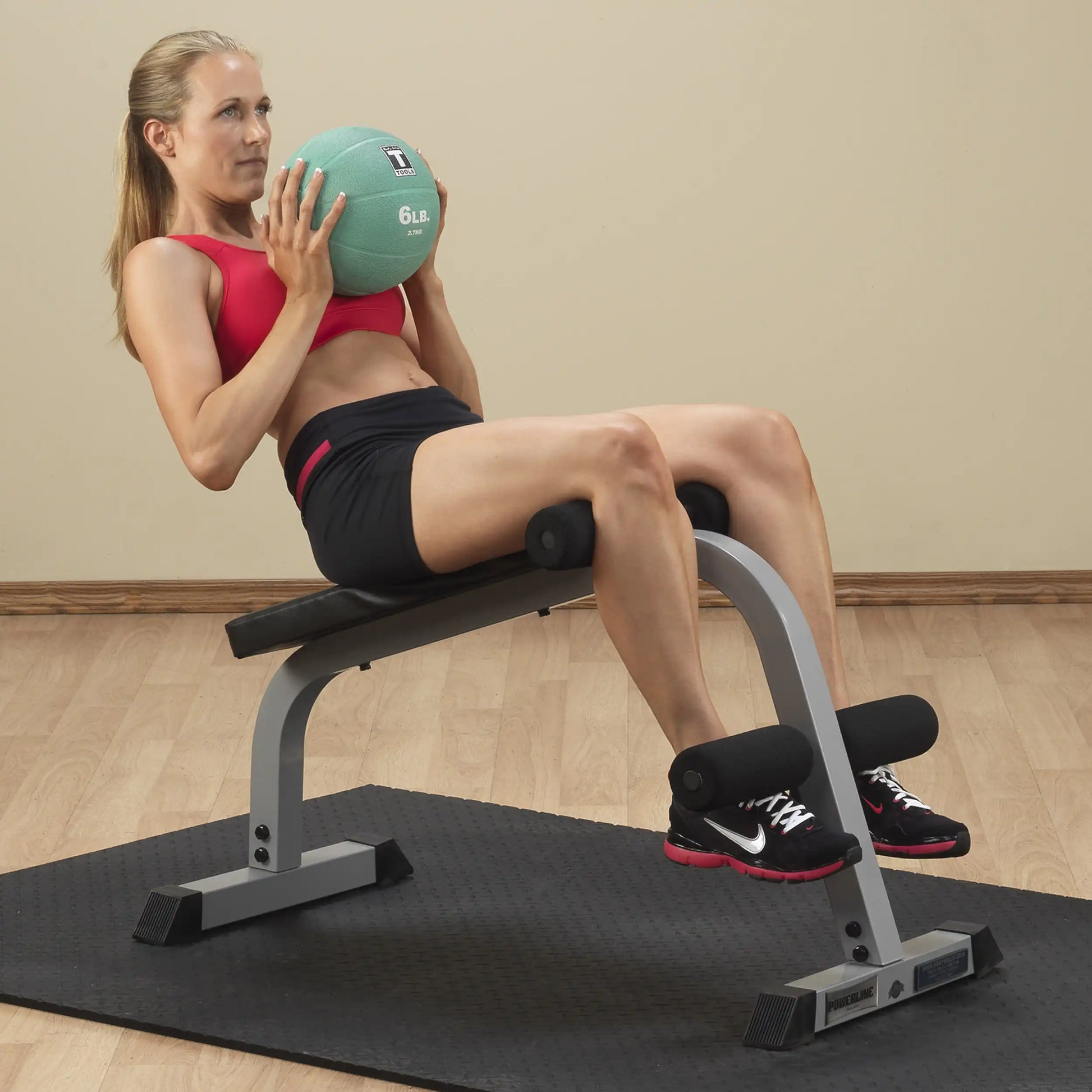 A woman in a red sports bra and black shorts sits on a workout bench, holding a light green medicine ball close to her chest while performing an exercise.