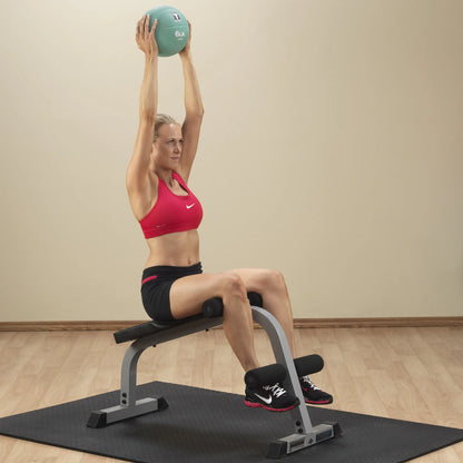 A woman in a red sports bra and black shorts is sitting on a workout bench, holding a medicine ball above her head with both hands, demonstrating a strength training exercise. The background features a neutral-colored wall and a rubber exercise mat on the floor.