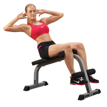 A woman in a pink sports bra and black shorts performing an abdominal exercise on a workout bench, with her hands behind her head and her legs secured in place.