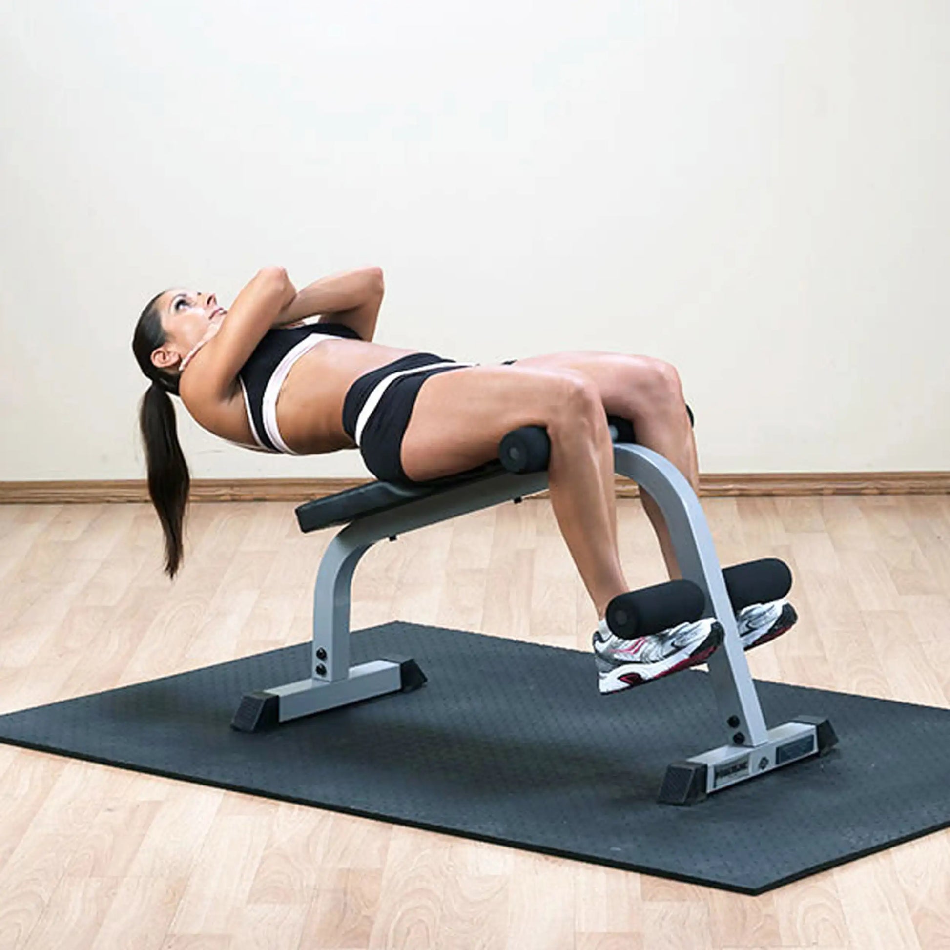 A woman performing an exercise on a bench, lying back with her arms crossed, wearing a sports bra and shorts, on a gym mat.