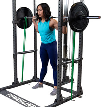 A person performing a squat with a barbell on their shoulders inside a power rack, wearing a blue tank top and navy leggings, with green resistance bands attached to the rack.