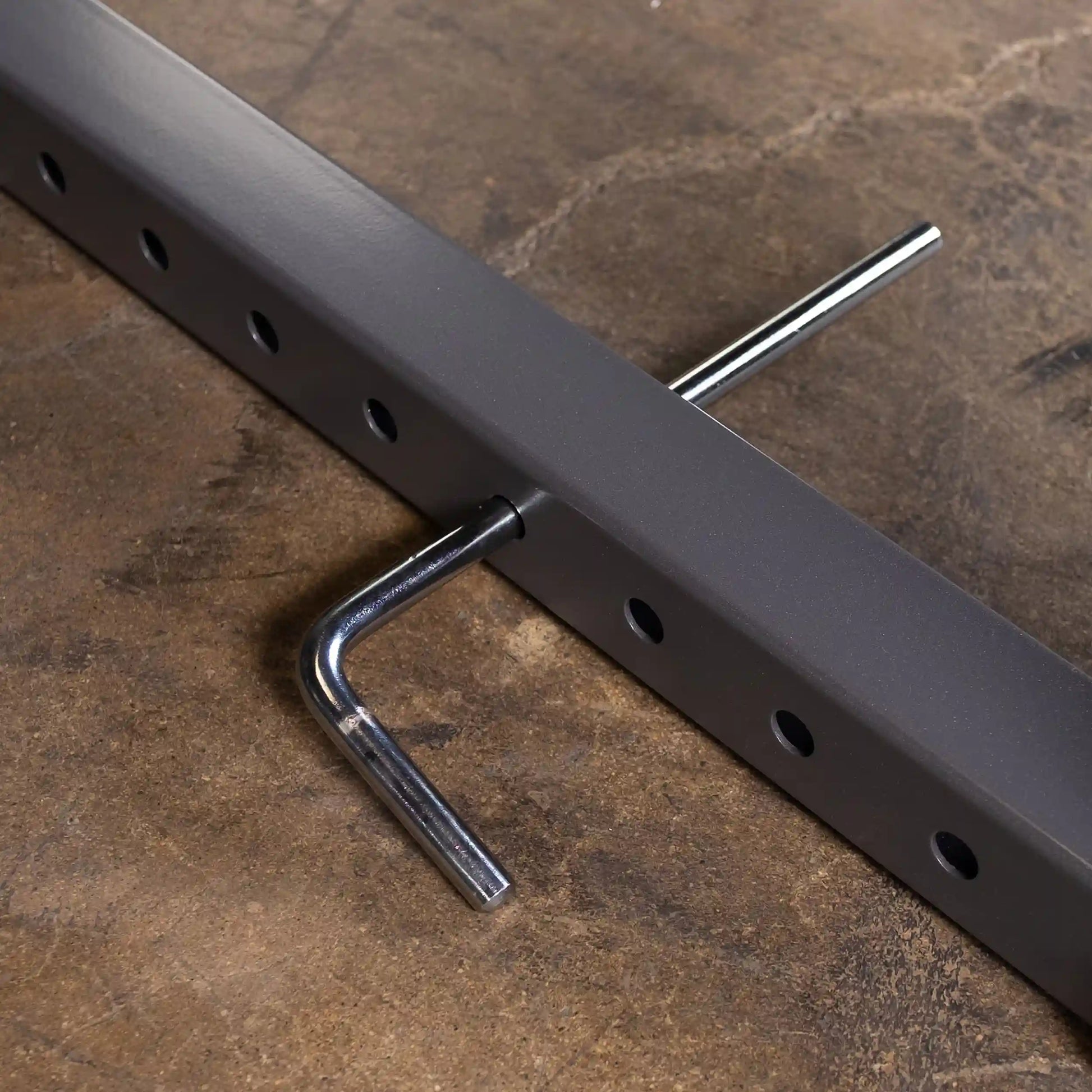 Close-up of a gray metal bar with multiple holes and a chrome hook attached, resting on a textured surface.