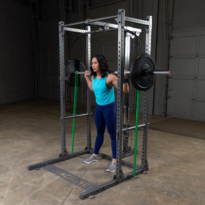 A woman performing a squat with a barbell on her shoulders inside a gym, wearing a turquoise tank top and blue leggings, using a squat rack with resistance bands attached.