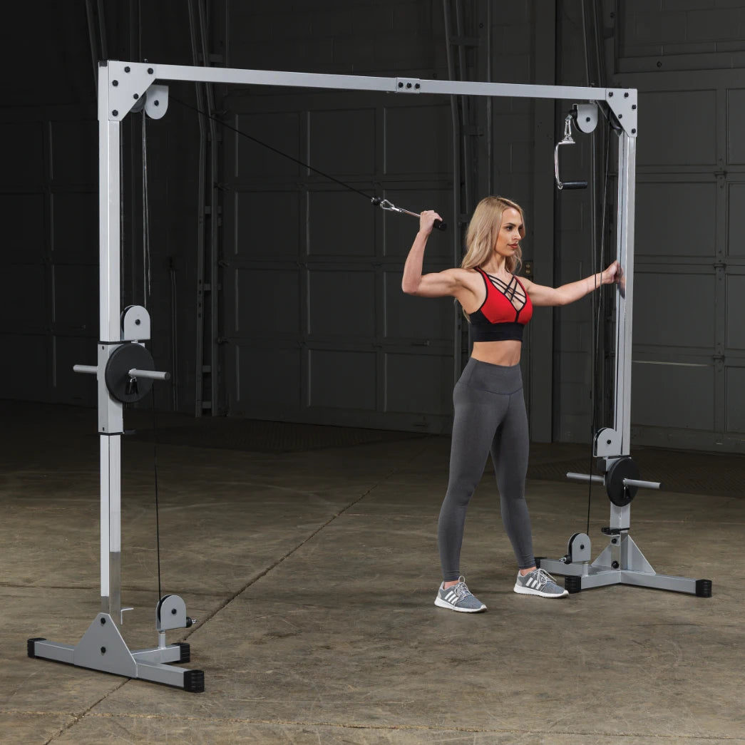 A woman in a red sports bra and gray leggings is performing an exercise using a cable machine in a gym setting. She is pulling the cable with one arm while maintaining a strong stance. The background features a concrete floor and a garage-style wall.