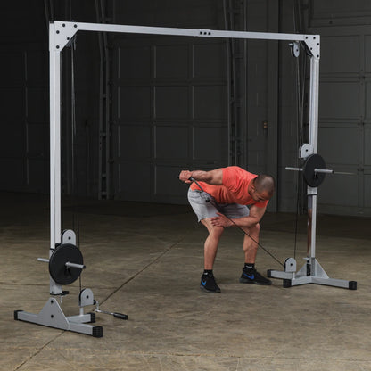 A person in a red shirt and gray shorts is performing a cable exercise using a dual pulley machine in a gym setting. The individual is bent slightly forward, gripping the cable handle with one hand while adjusting their stance.