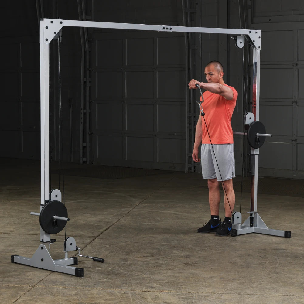 A man in a bright orange shirt and gray shorts stands in a gym setting, preparing to perform an exercise using a cable machine.