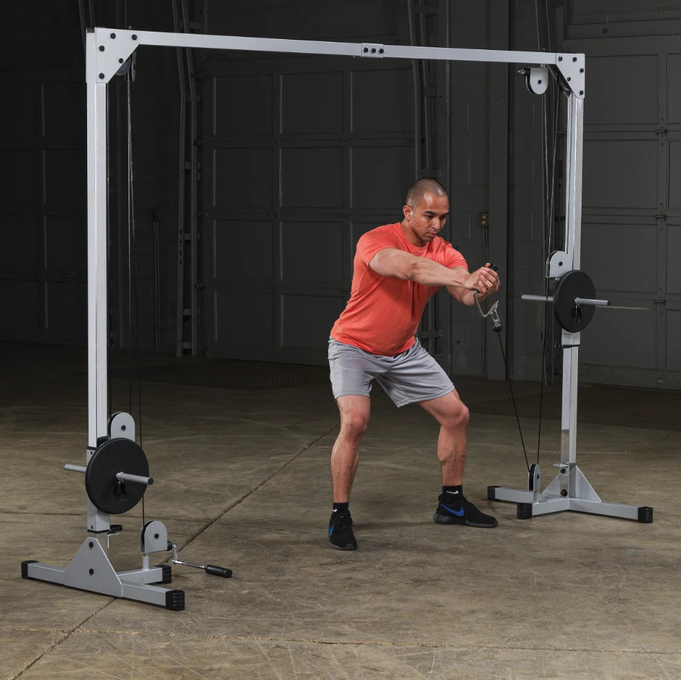 A man in an orange shirt and gray shorts performs a cable exercise using a dual pulley machine in a gym setting. The background shows a spacious area with a concrete floor and garage-style doors.