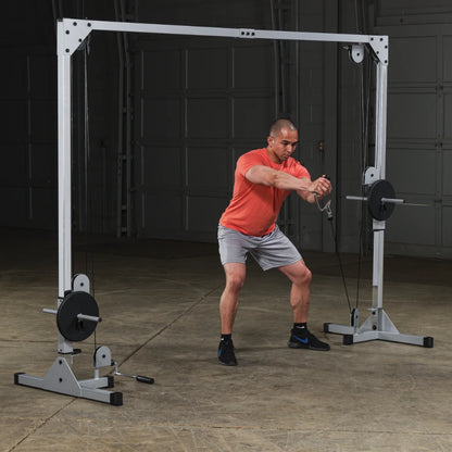 A man in an orange shirt and gray shorts performs a cable exercise using a dual pulley machine in a gym setting. The background shows a spacious area with a concrete floor and garage-style doors.