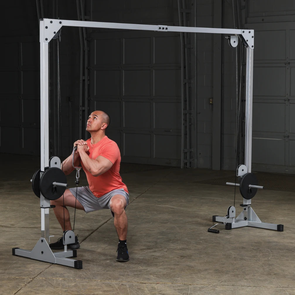A man in an orange shirt and gray shorts is performing a squat exercise using a cable machine in a gym setting. The background features a large garage door and concrete flooring.
