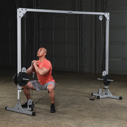 A man in an orange shirt and gray shorts is performing a squat exercise using a cable machine in a gym setting. The background features a large garage door and concrete flooring.