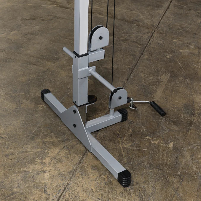 Close-up view of a gym cable machine with pulleys and a foot pedal on a textured floor.
