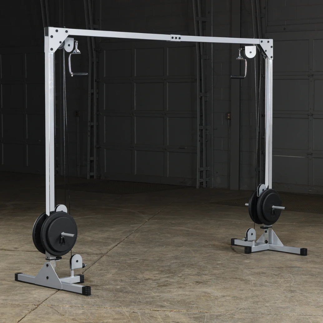 A weight training machine with a white frame, featuring two pulleys on either side and weights on the base, set in a spacious gym environment with a concrete floor and dim lighting.