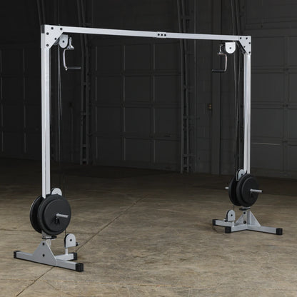A weight training machine with a white frame, featuring two pulleys on either side and weights on the base, set in a spacious gym environment with a concrete floor and dim lighting.