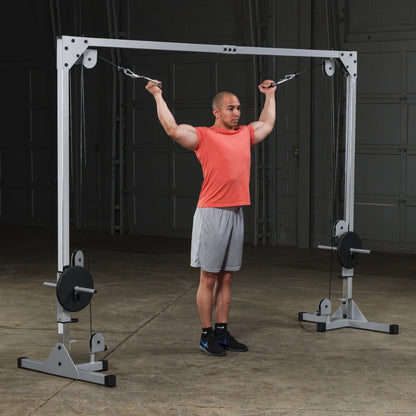 A man in a red shirt and gray shorts performs a cable exercise with both arms extended above his head, using a cable machine in a gym setting.