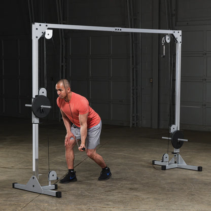 A man in a red t-shirt and gray shorts performs a cable exercise using a multi-station gym equipment in a spacious gym setting.