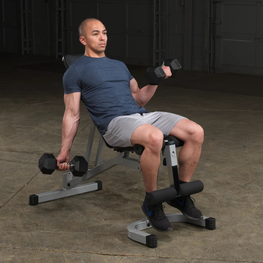 A person seated on a workout bench, performing a dumbbell exercise with weights in both hands, wearing a blue shirt and gray shorts, in a gym setting with a concrete floor.