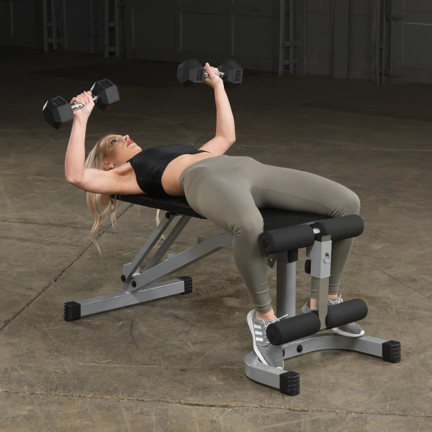 A woman lying on a weight bench, performing a dumbbell exercise with free weights in both hands, wearing a black crop top and olive green leggings, in a gym setting.