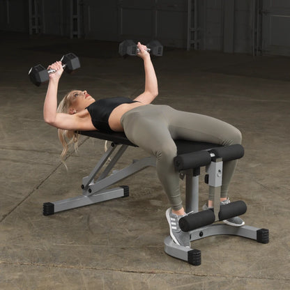 A woman lying on a weight bench, performing a dumbbell exercise with free weights in both hands, wearing a black crop top and olive green leggings, in a gym setting.