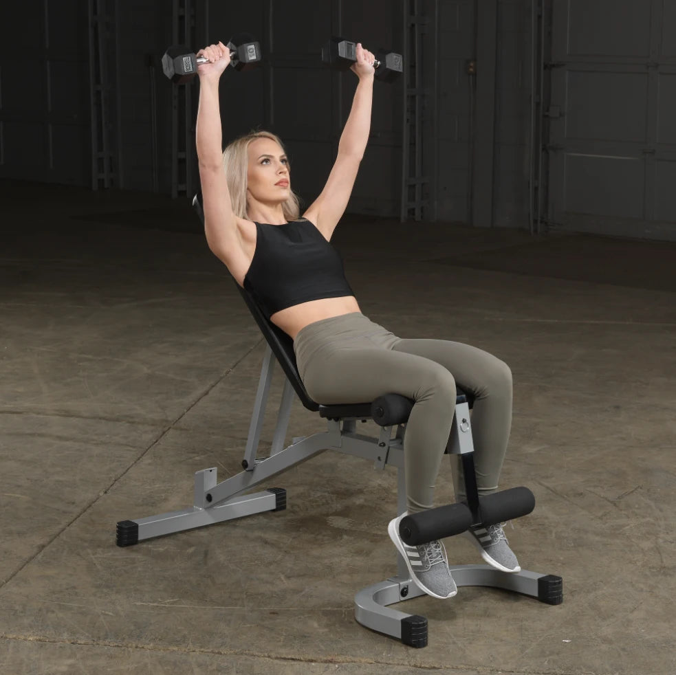 A woman with blonde hair is performing a dumbbell press while seated on a weight bench. She is wearing a black crop top and olive green leggings, lifting weights overhead in a gym setting.