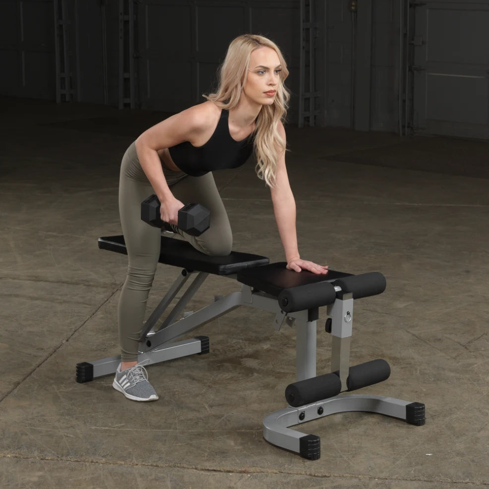 A woman with long blonde hair is performing a dumbbell exercise on a bench in a gym setting. She is leaning slightly forward, holding a dumbbell in her right hand, while her left hand rests on the bench. She is wearing a black tank top and olive green leggings.