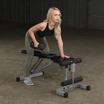 A woman with long blonde hair is performing a dumbbell exercise on a bench in a gym setting. She is leaning slightly forward, holding a dumbbell in her right hand, while her left hand rests on the bench. She is wearing a black tank top and olive green leggings.