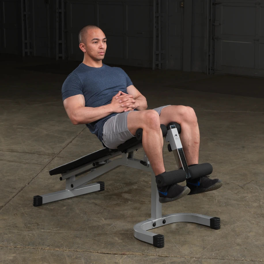 A man sitting on a workout bench with his hands clasped, wearing a blue t-shirt and gray shorts, preparing for an exercise session in a gym setting.