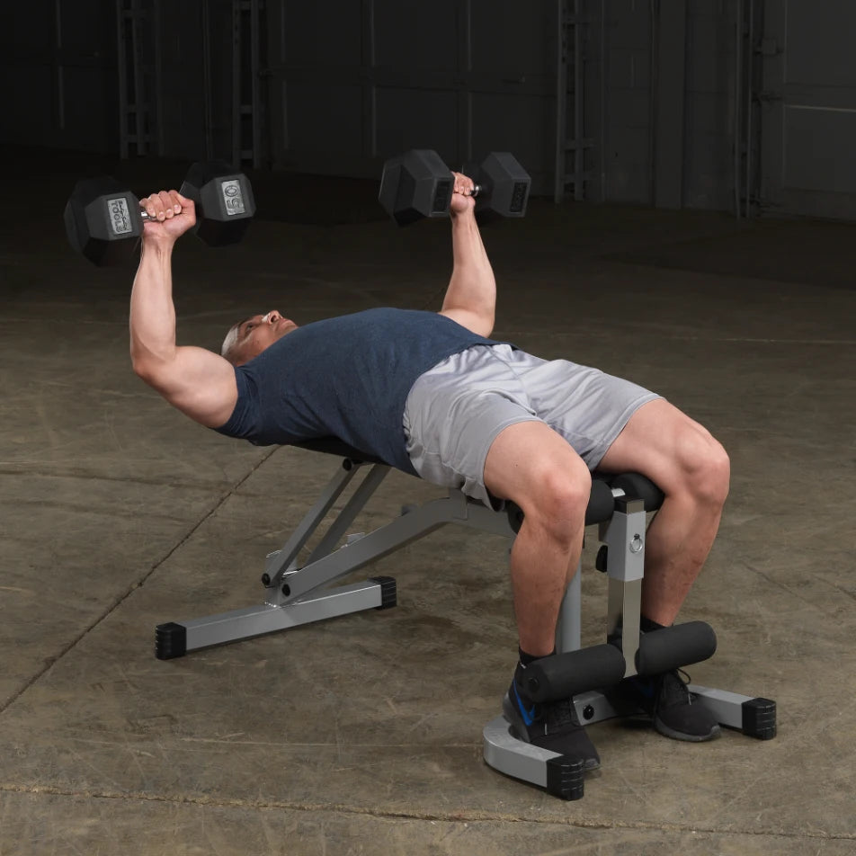 A man lying on a bench press, lifting dumbbells above his chest in a gym setting. He is wearing a blue shirt and gray shorts, with his feet secured on a leg holder. The gym has a concrete floor and a simple, industrial backdrop.