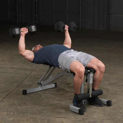A man lying on a bench press, lifting dumbbells above his chest in a gym setting. He is wearing a blue shirt and gray shorts, with his feet secured on a leg holder. The gym has a concrete floor and a simple, industrial backdrop.