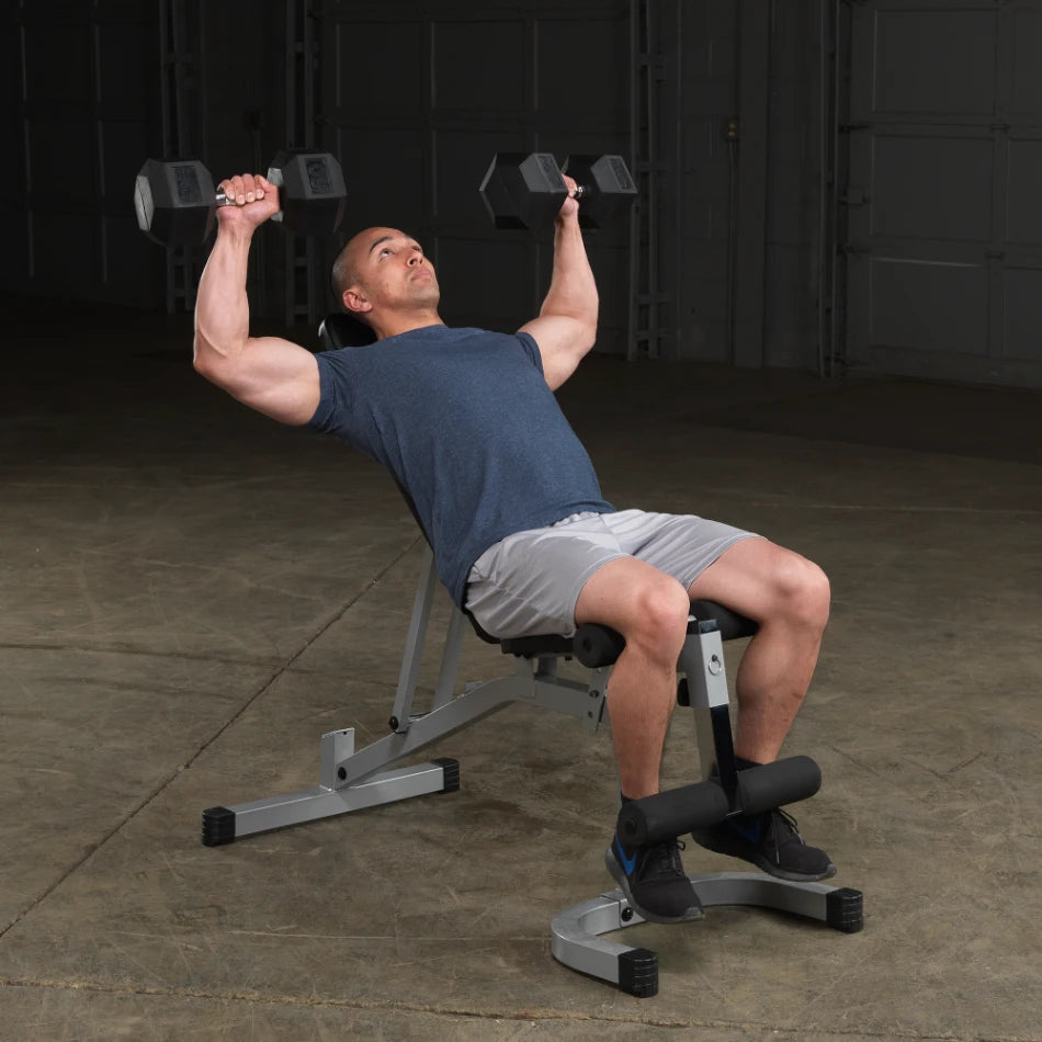 A man performing a dumbbell shoulder press exercise while seated on a weight bench in a gym setting, wearing a blue t-shirt and gray shorts.