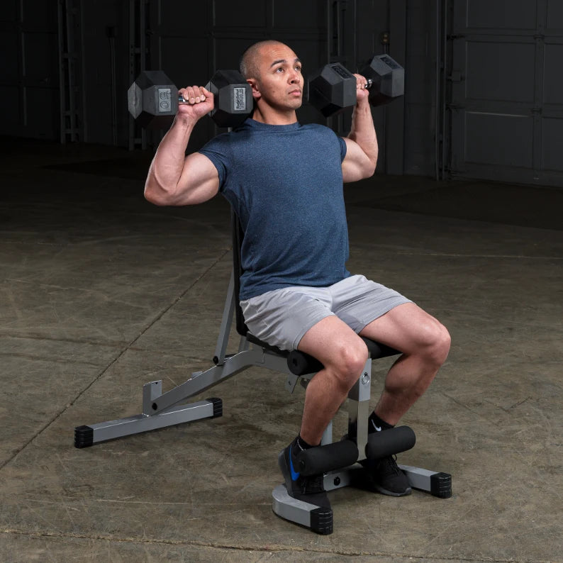 A man seated on a weight bench performing a shoulder press with dumbbells, wearing a blue shirt and gray shorts, in a gym setting.