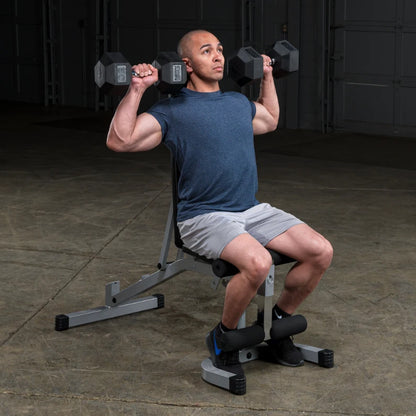 A man seated on a weight bench performing a shoulder press with dumbbells, wearing a blue shirt and gray shorts, in a gym setting.