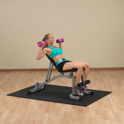 A woman in a turquoise sports bra and black shorts performs a shoulder press with pink dumbbells while seated on an adjustable bench in a gym setting.