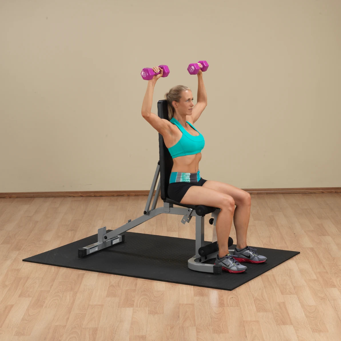 A woman in a turquoise sports bra and black shorts is seated on a weight bench, performing a shoulder press with pink dumbbells raised above her head. The setting features a light-colored wall and wooden flooring.
