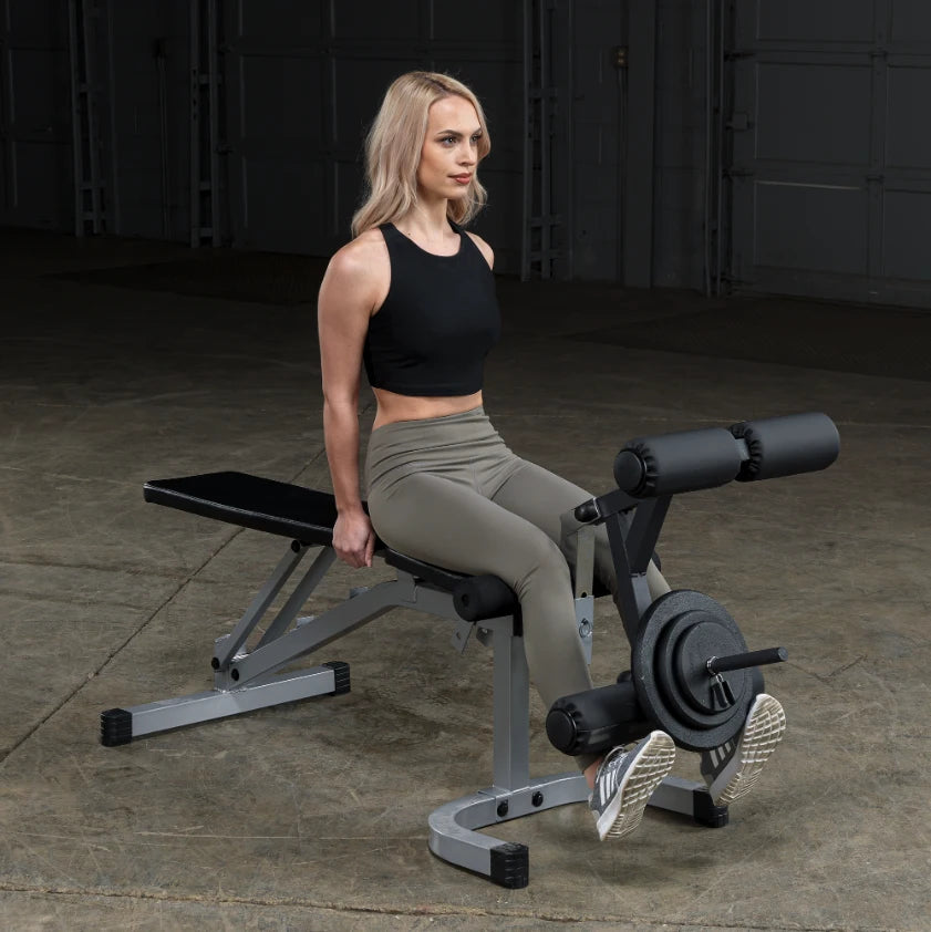 A woman with long blonde hair is sitting on a weight bench, preparing to perform an exercise. She is wearing a black crop top and olive green leggings, with her feet on the equipment's footrest. The background is a dimly lit gym space.