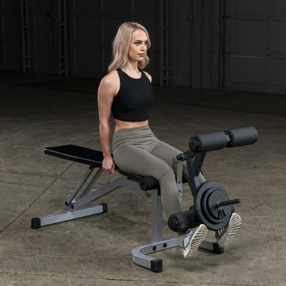 A woman with long blonde hair is sitting on a weight bench, preparing to perform an exercise. She is wearing a black crop top and olive green leggings, with her feet on the equipment's footrest. The background is a dimly lit gym space.