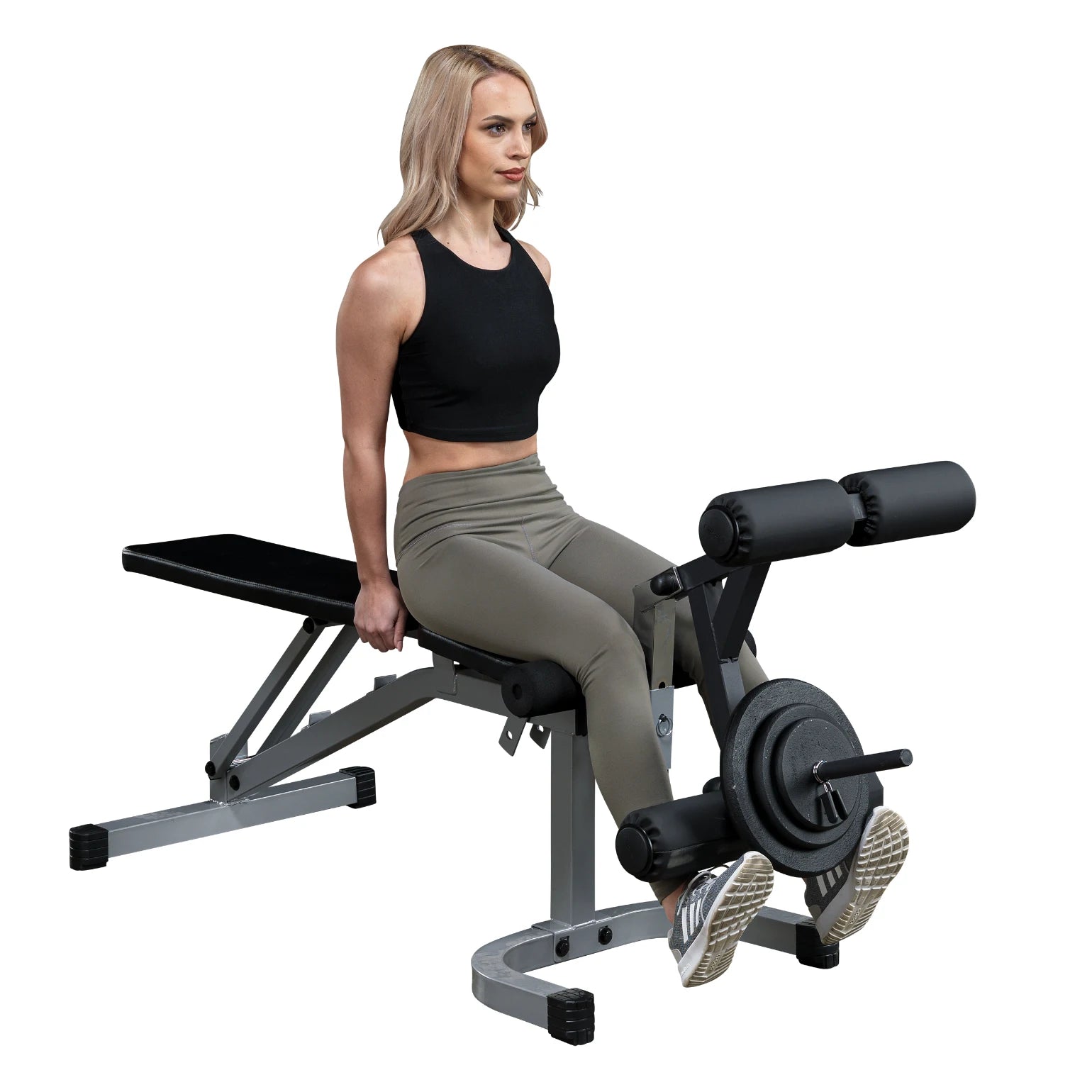 A woman with long blonde hair is sitting on a weight bench, preparing to perform a leg exercise. She is wearing a black crop top and olive green leggings, with her feet positioned on the exercise equipment that has weights attached.