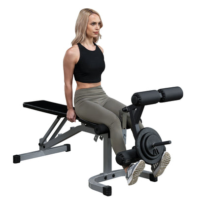 A woman with long blonde hair is sitting on a weight bench, preparing to perform a leg exercise. She is wearing a black crop top and olive green leggings, with her feet positioned on the exercise equipment that has weights attached.