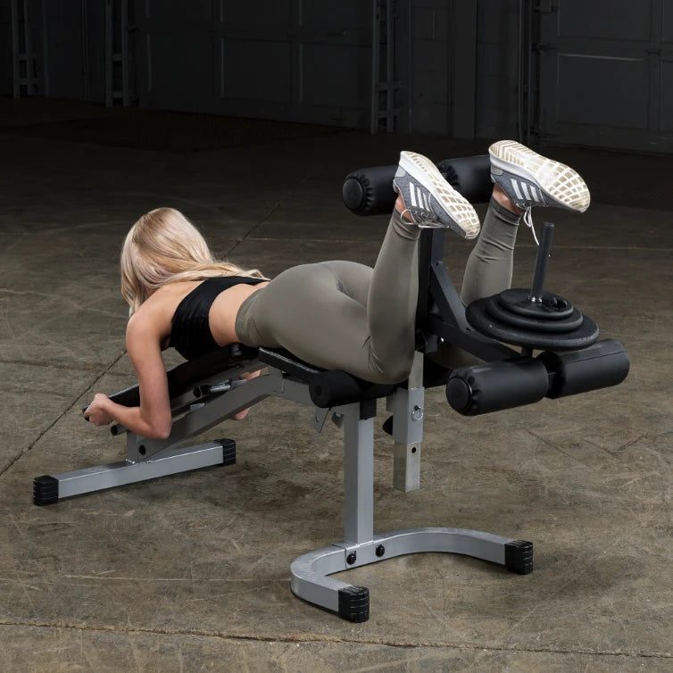 A person performing an exercise on a leg curl machine in a gym setting, wearing a black top and olive green leggings, with weights on the machine.