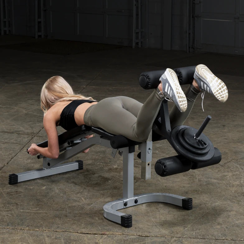 A person performing a workout on a gym machine designed for leg exercises, wearing a black crop top and olive green leggings, with a focus on their form and technique in a spacious gym environment.