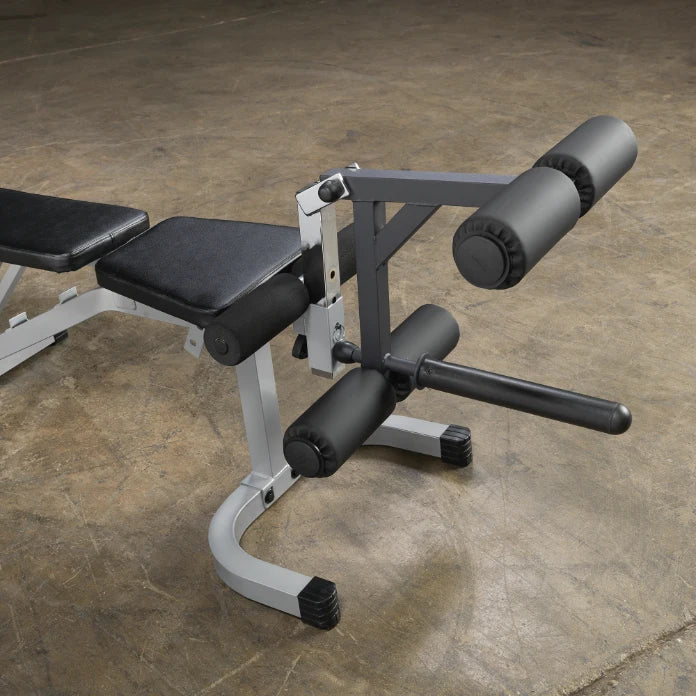 A close-up view of a gym equipment bench designed for leg exercises, featuring padded rollers and a black and silver frame, set against a concrete floor.