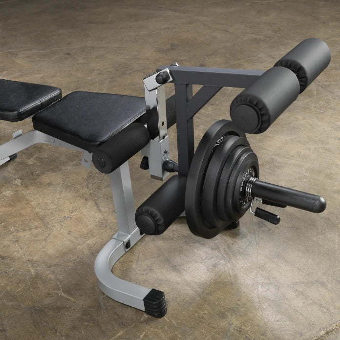 A close-up view of a weight bench with a leg extension attachment, featuring padded rollers and a weight plate on the side, set against a concrete floor.