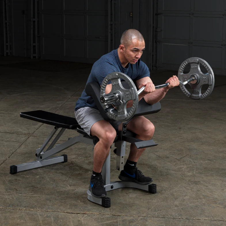 A man with a shaved head is seated on a weight bench, performing a bicep curl with a barbell loaded with weights. He wears a blue t-shirt and gray shorts, focusing intently on his workout in a gym setting.