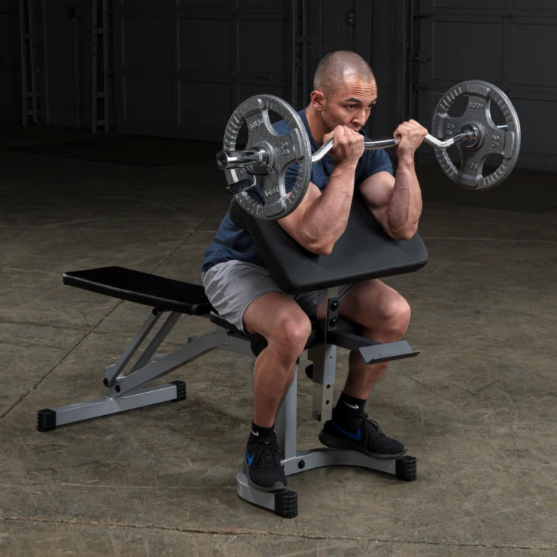 A person with short hair is performing a bicep curl with a barbell while seated on a weight bench in a gym setting. The individual is focused and lifting weights, showcasing strength training.