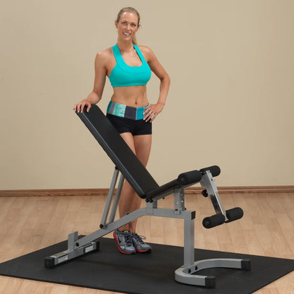 A woman in a turquoise sports bra and black shorts stands confidently next to an adjustable workout bench on a black exercise mat, smiling at the camera.
