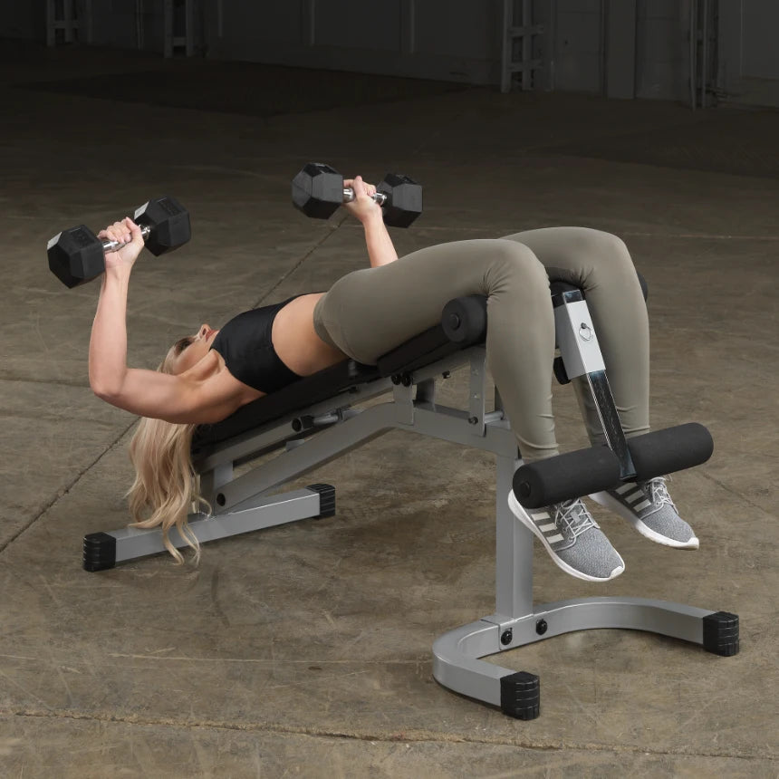 A woman lying on a weight bench performs a dumbbell chest press, holding a dumbbell in each hand. She is wearing a black sports top and olive green leggings, with her long hair down. The setting appears to be a gym or workout space.